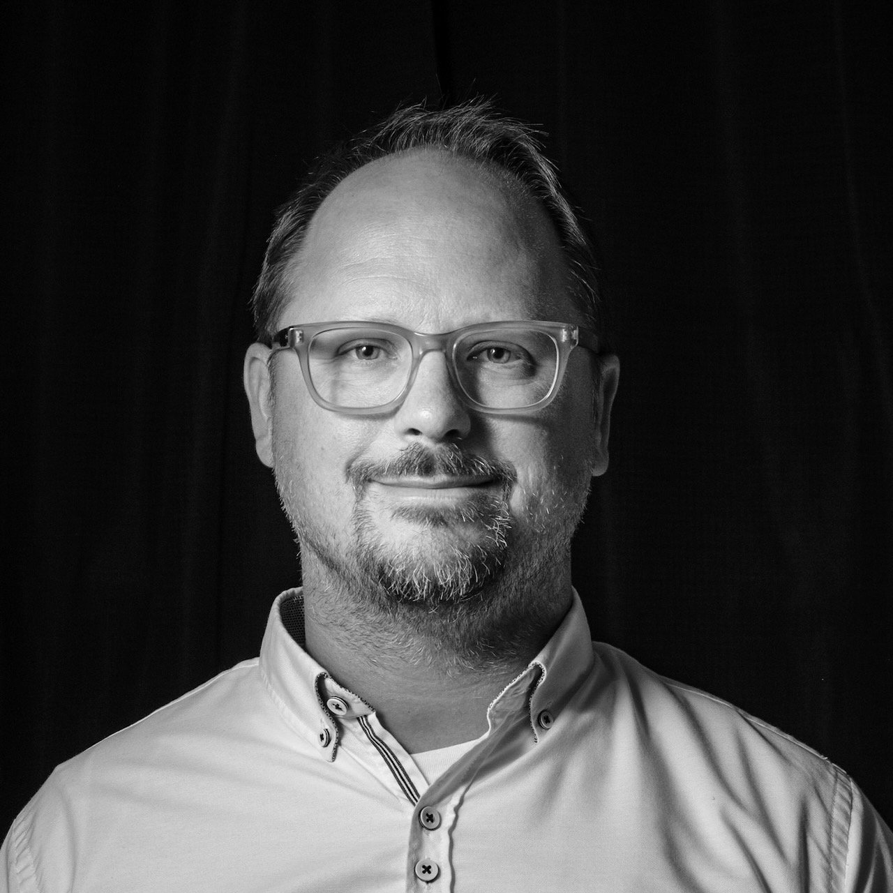 Black and white portrait of a man with glasses and a beard wearing a collared shirt, smiling gently.